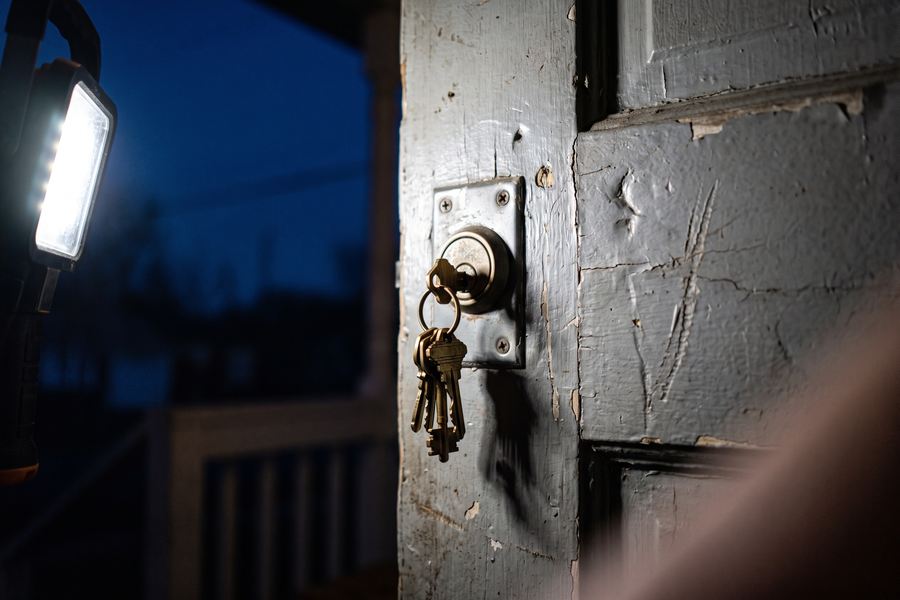 House keys left in a front door lock, lit by a handheld work light at twilight